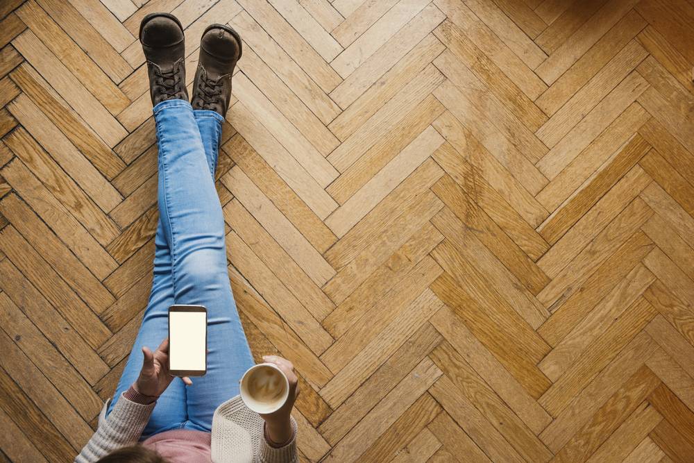 Ženska z mobilnim telefonom pije kavo in sedi na lesenih tleh, ki so v obliki ribje kosti.