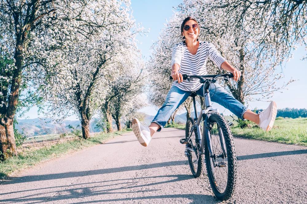 Nasmejana ženska s treking kolesom kolesari po stezici, obdani s cvetočimi drevesi.