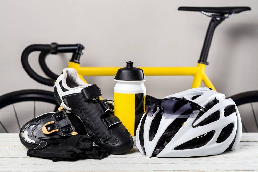 Specializirana kolesarska oblačila in oprema - čelada, očala, rokavice in kolesarski čevlji s cestnim kolesom v ozadju.
