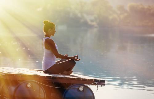 Pogled na žensko, ki sedi ob jezeru in se sprošča z meditacijo.