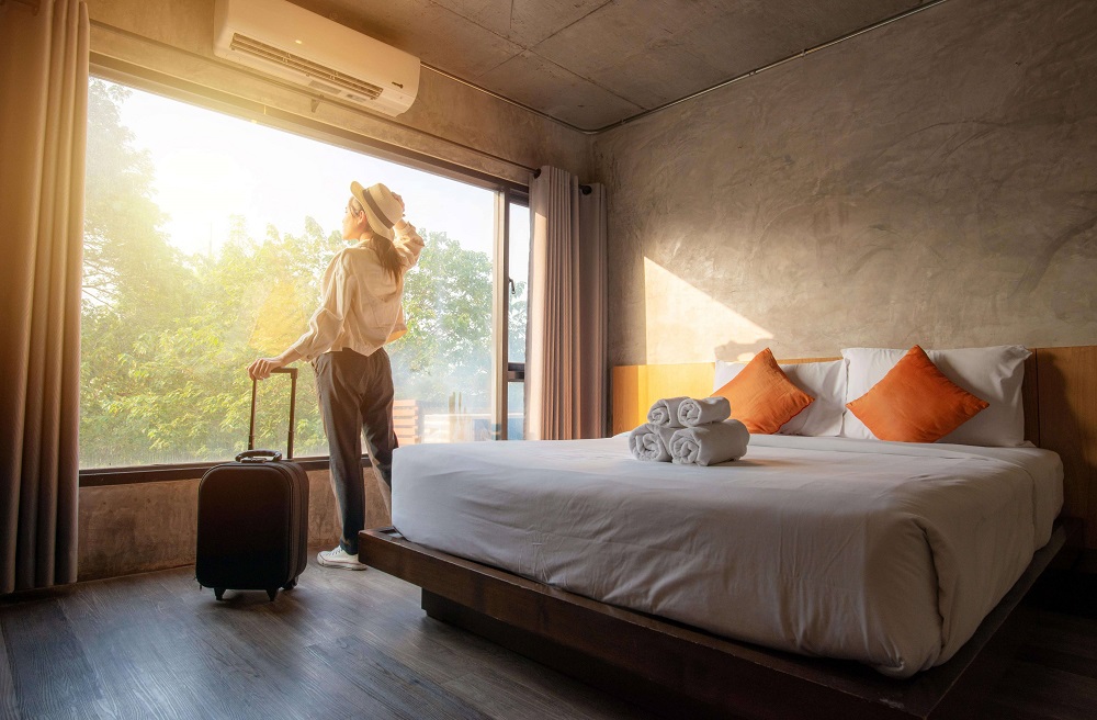 Ženska s potovalnim kovčkom stoji poleg postlane postelje in zre skozi veliko hotelsko okno.